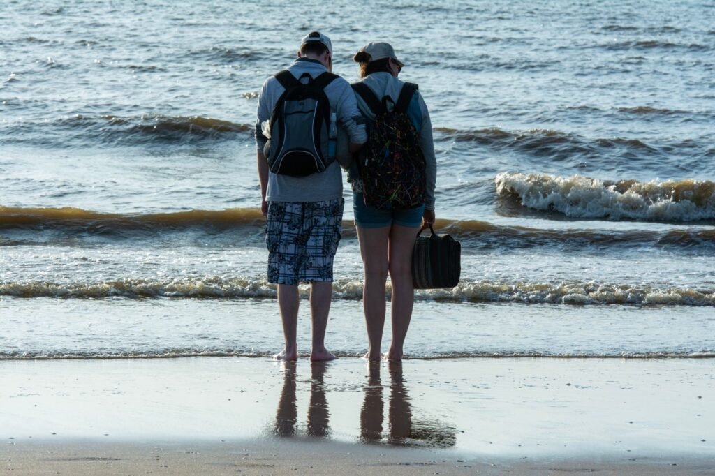 A couple stands barefoot on the beach facing the ocean with backpacks, symbolizing relaxation and adventure.