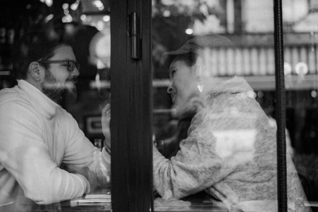 Black and white photo of a couple having a romantic moment in a cafe, seen through a window.