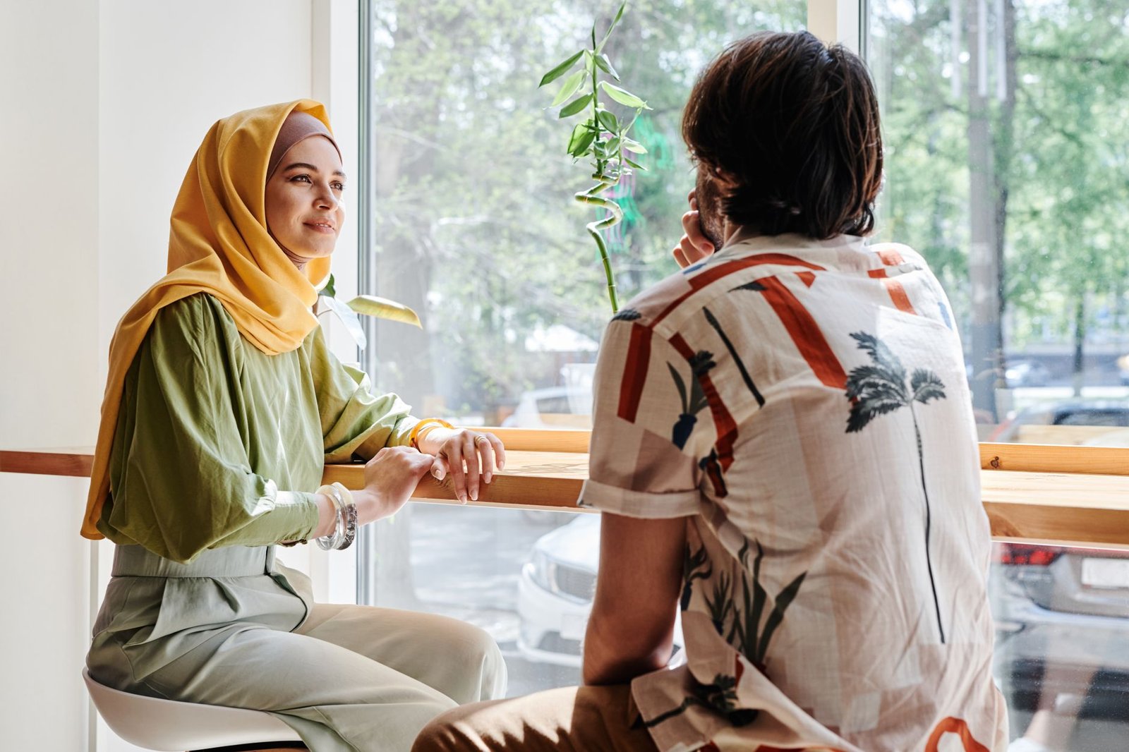 A couple enjoys a relaxed conversation at a bright café with leafy views.