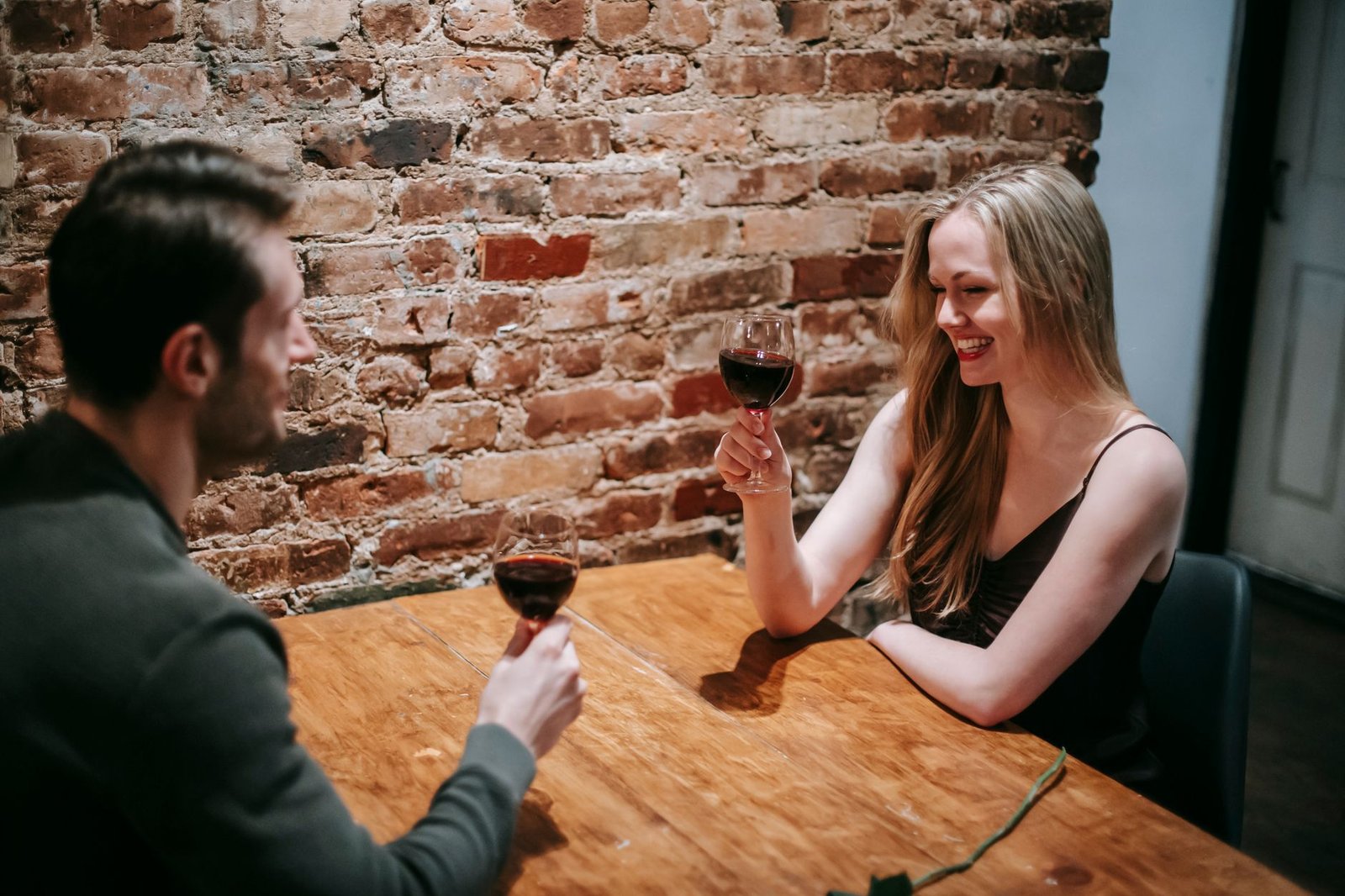 Side view of young smiling couple in elegant clothes sitting in cafeteria at table near brick wall while having romantic dinn